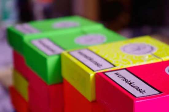 In neon Farben gestapelte Schachteln. Oben drauf der schwarze Schriftzug Wunstkunst auf weißem Grund.