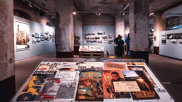 Eine Ausstellungshalle in einem alten Gebäude mit rauen, steinernen Säulen. Im Vordergrund werden Magazine und Bücher auf einer Vitrine präsentiert.