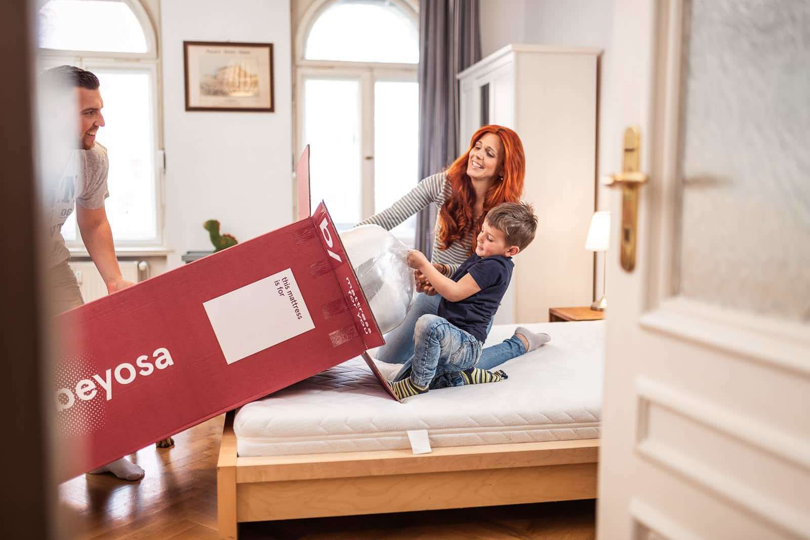 Eine lachende Frau mit roten Haaren und ein Kind beim Auspacken einer weißen Matratze aus einem großen roten Karton der Marke