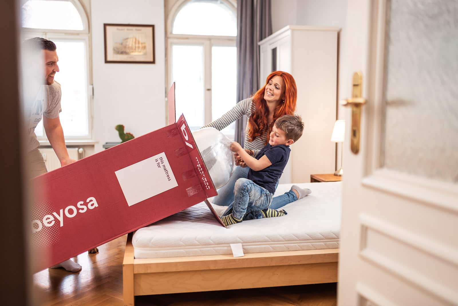 Eine lachende Frau mit roten Haaren und ein Kind beim Auspacken einer weißen Matratze aus einem großen roten Karton der Marke