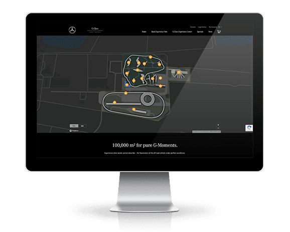Ein Desktop-Monitor zeigt eine interaktive Gel&auml;ndekarte des &bdquo;G-Class Experience Center&ldquo;. Auf einem dunklen Hintergrund sind die verschiedenen Teststrecken und Fahrtrainings-Areale mit orangefarbenen Standort-Pins markiert. Darunter steht der Slogan: &bdquo;100,000 m&sup2; for pure G-Moments.&ldquo;