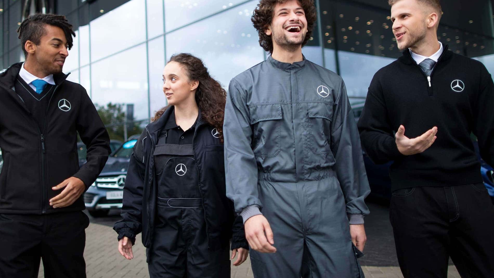Vier junge diverse Mercedesmitarbeiter:innen in unterschiedlicher Mercedes-Arbeitskleidung vor einer Mercedes-Niederlassung