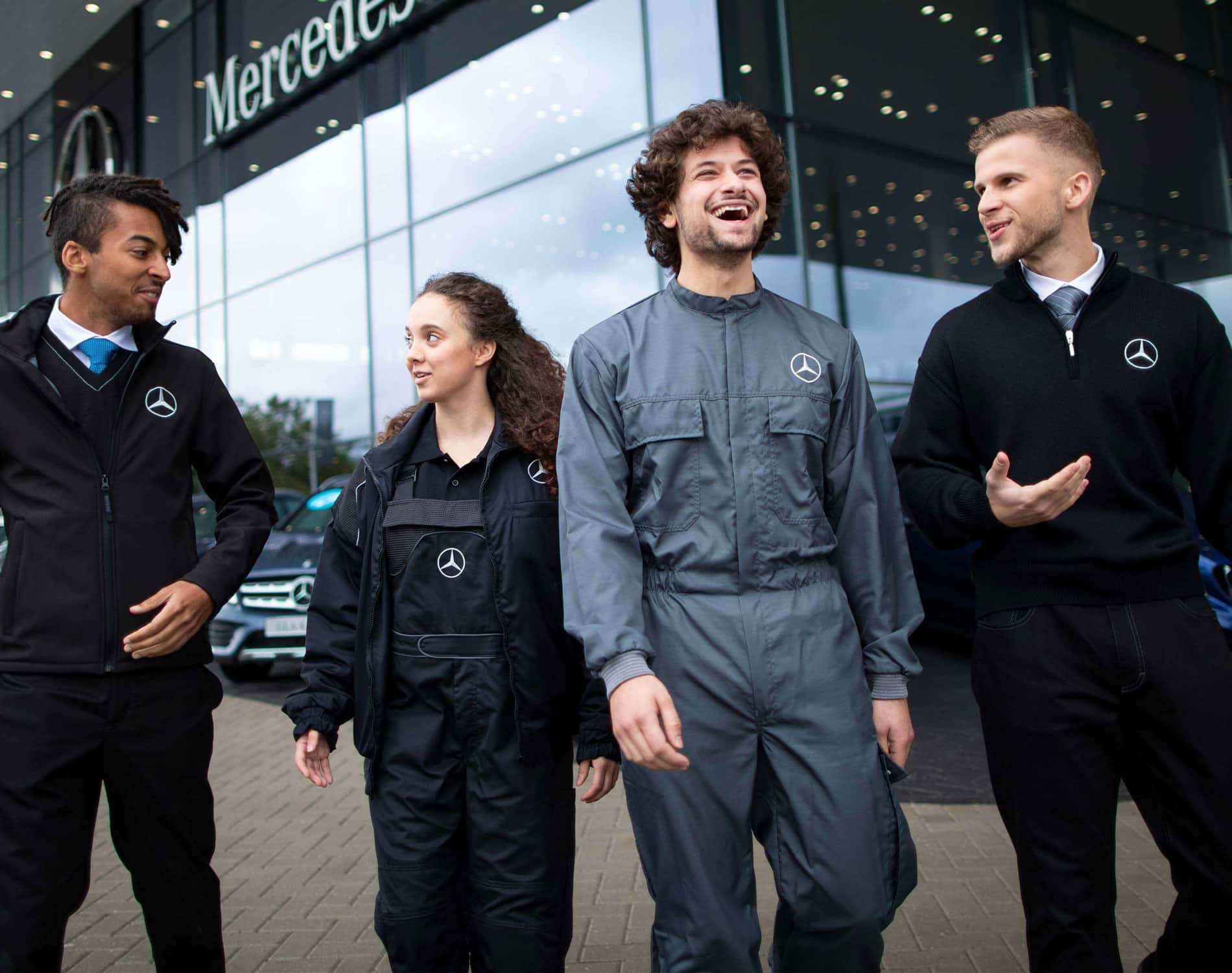 Vier junge diverse Mercedesmitarbeiter:innen in unterschiedlicher Mercedes-Arbeitskleidung vor einer Mercedes-Niederlassung