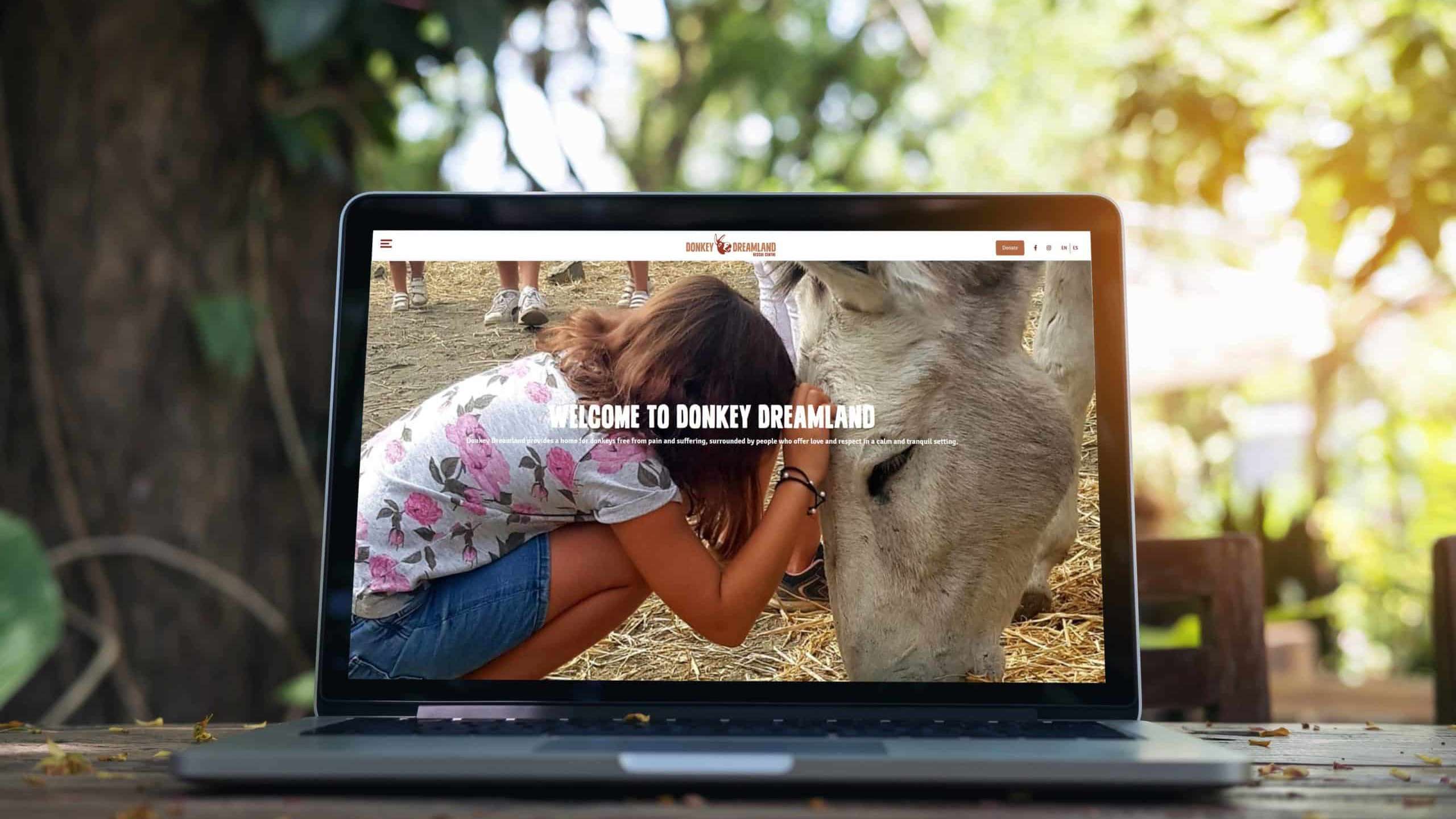 Schwarzer Laptop auf Holztisch mit der Website von Donkey Dreamland auf dem Bildschirm. Im Hintergrund Bäume.
