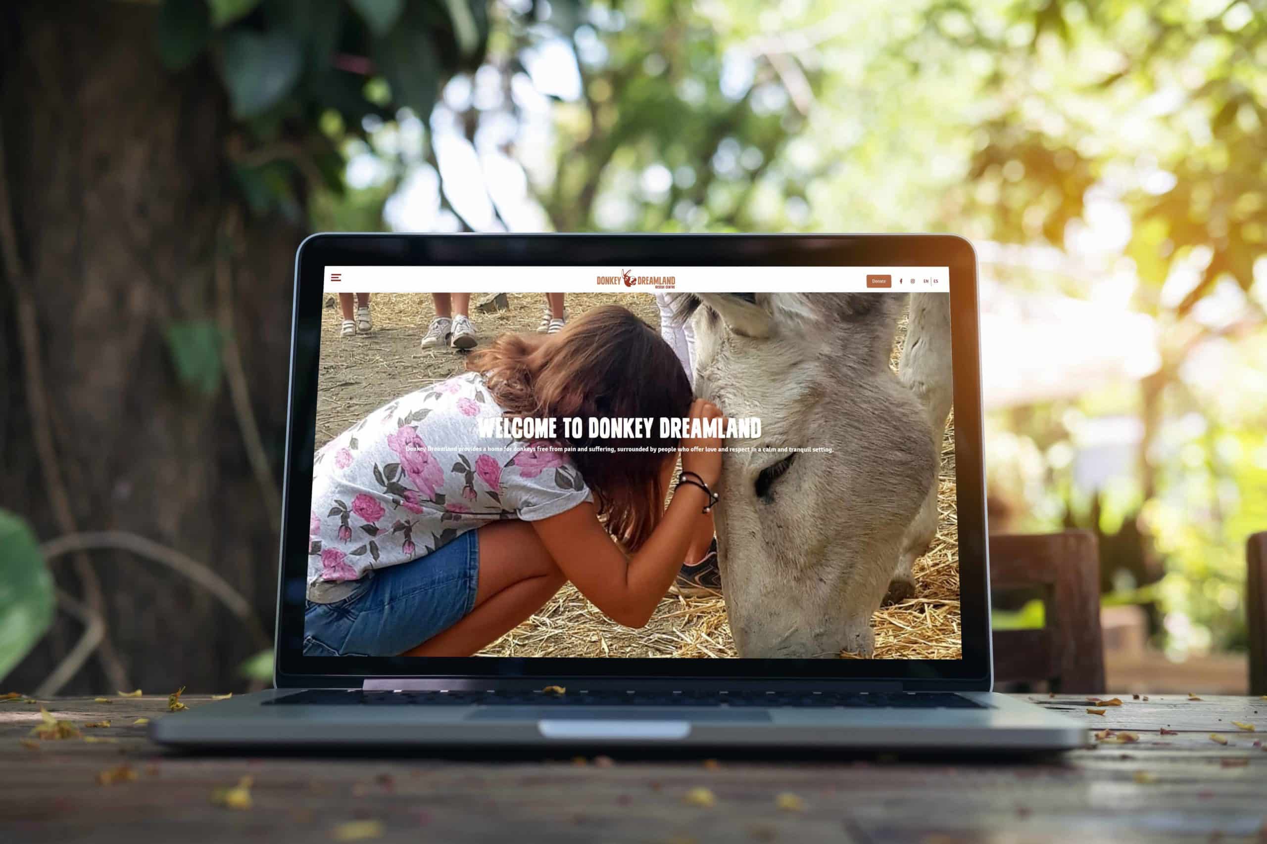 Schwarzer Laptop auf Holztisch mit der Website von Donkey Dreamland auf dem Bildschirm. Im Hintergrund Bäume.