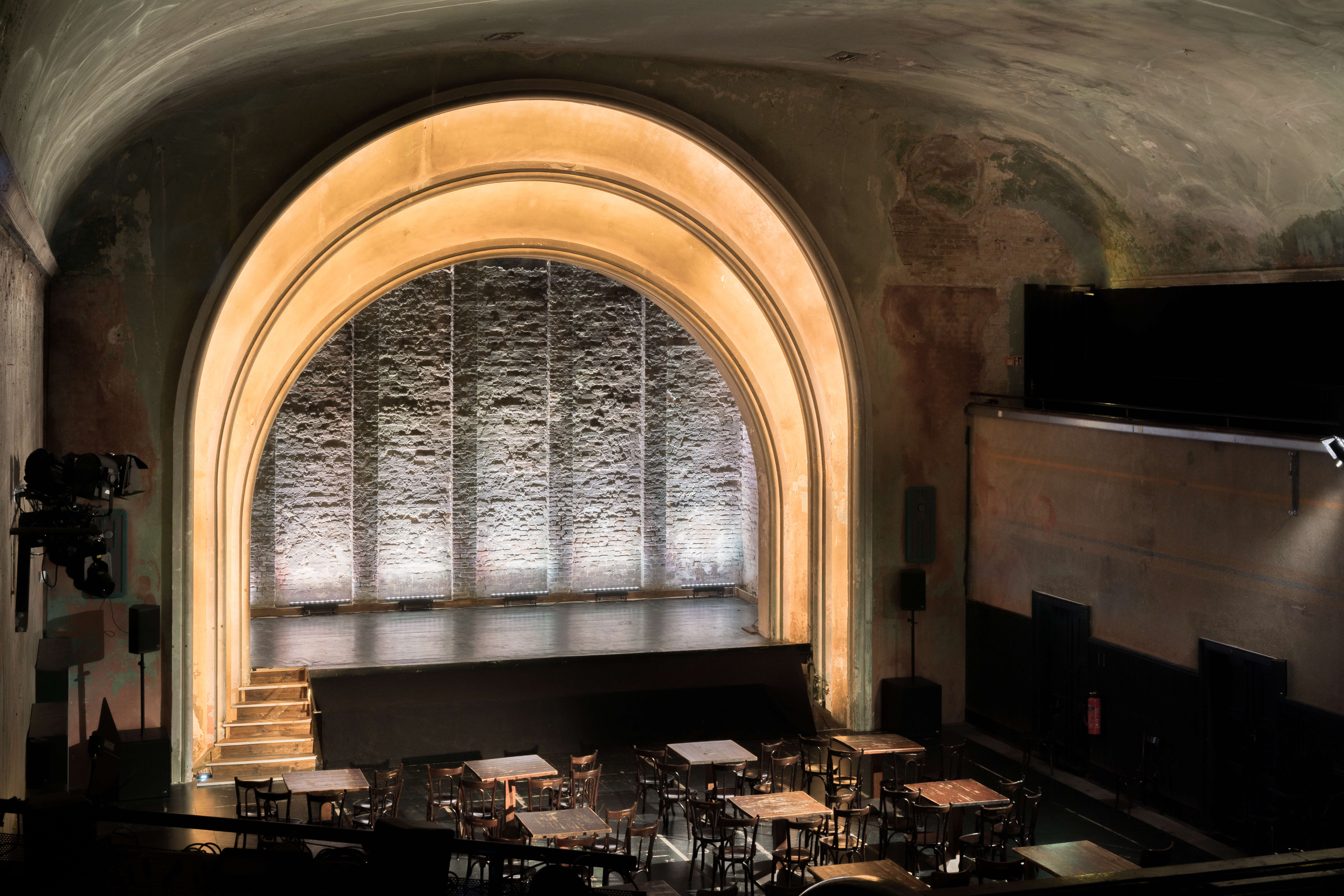 Innenansicht des „Theater im Delphi“ in Berlin. Der Saal besticht durch seinen charmanten Industrie-Stil mit hohen Gewölbedecken und unverputzten Wänden. Im Mittelpunkt steht eine Bühne unter einem großen, beleuchteten Rundbogen, davor sind Holztische und Stühle wie in einem Varieté angeordnet.