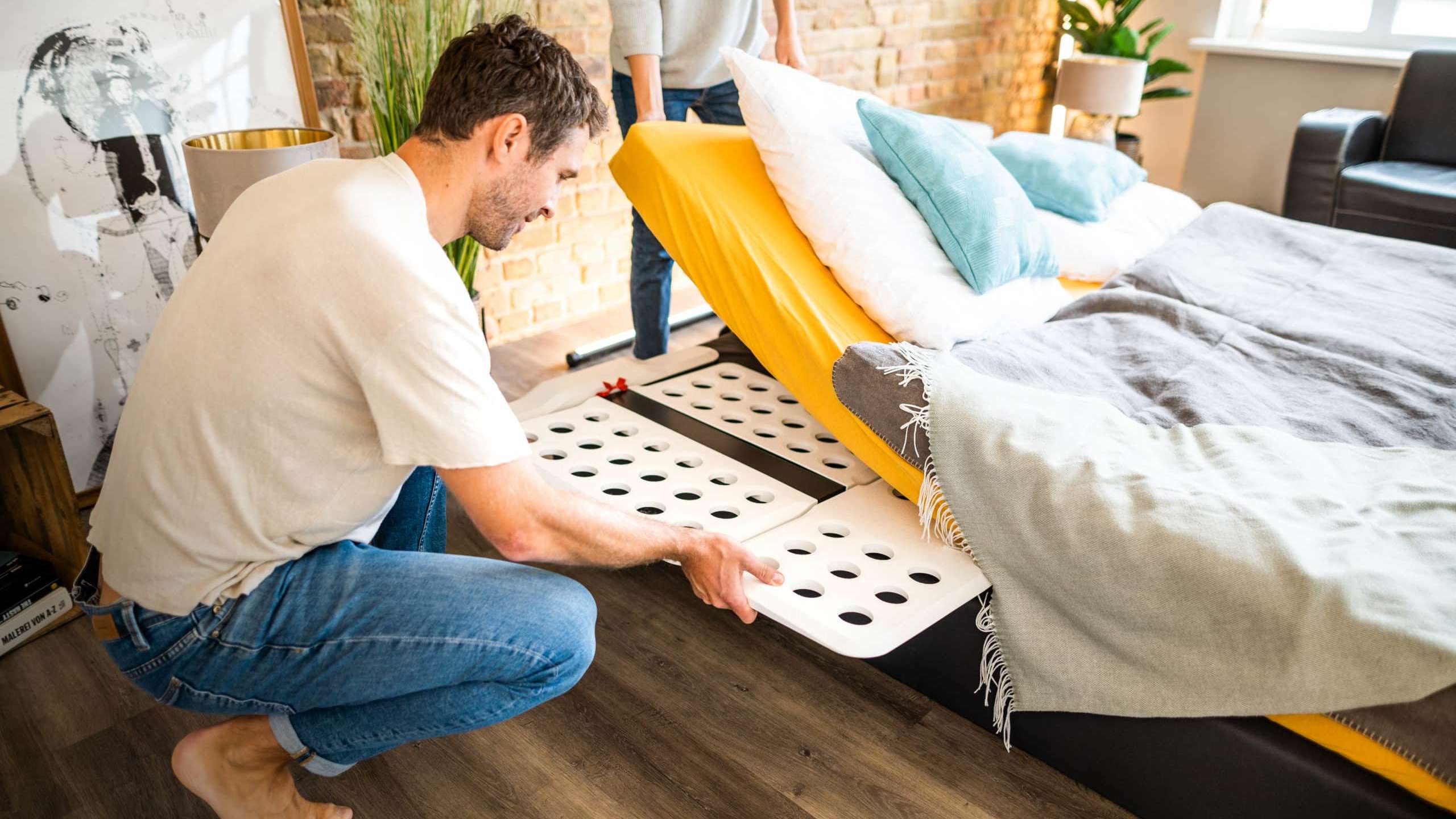 Ein Mann kniet barfuß auf einem Holzboden und montiert oder justiert ein weißes, modulares Bettsystem mit runden Aussparungen. Im Hintergrund hält eine weitere Person eine Matratze hoch, die auf einem gelben Spannbettlaken liegt. Das Schlafzimmer hat eine Backsteinwand und ist modern eingerichtet.