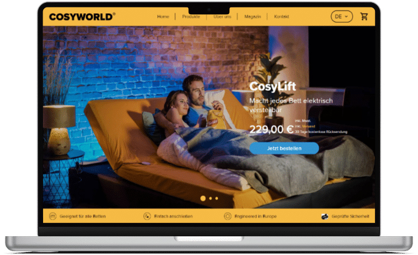 Ein Laptop zeigt die Startseite von CosyWorld. Zu sehen ist ein Paar, das gem&uuml;tlich in einem verstellbaren Bett liegt, daneben der Produktname &bdquo;CosyLift&ldquo;, der Preis und ein blauer &bdquo;Jetzt bestellen&ldquo;-Button.