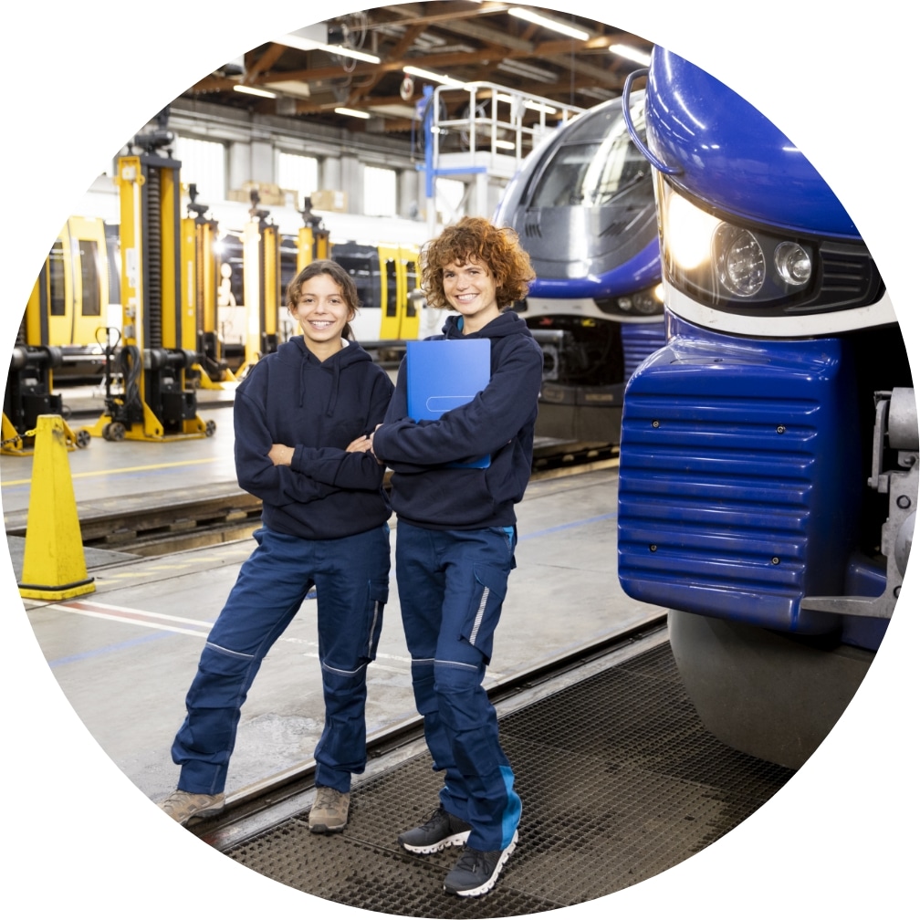 Zwei Frauen in Dunkelblauer Arbeitskleidung in einer Halle. Im Hintergrund blau/wei&szlig;e und Gelbe Bahnen/Z&uuml;ge.